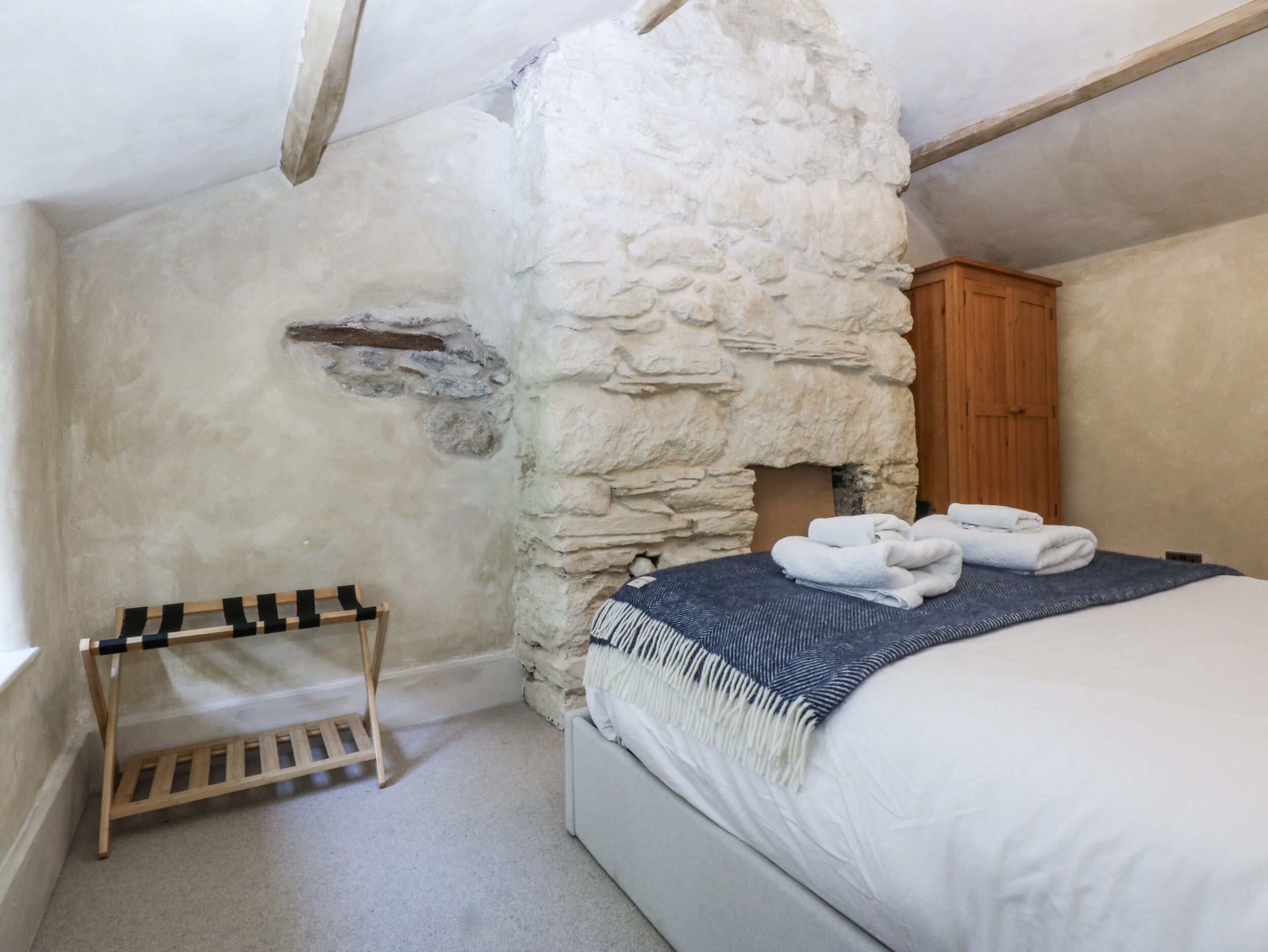 A bedroom with a stone wall, a wooden wardrobe, a bed with a blue and white striped blanket, and a wooden drying rack.