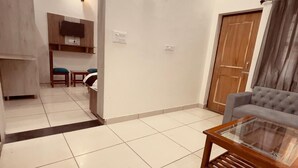 A spacious living area with a grey sofa, a glass-top coffee table, and a kitchen area in the background. - Vishram Guest House (Vikasnagar)