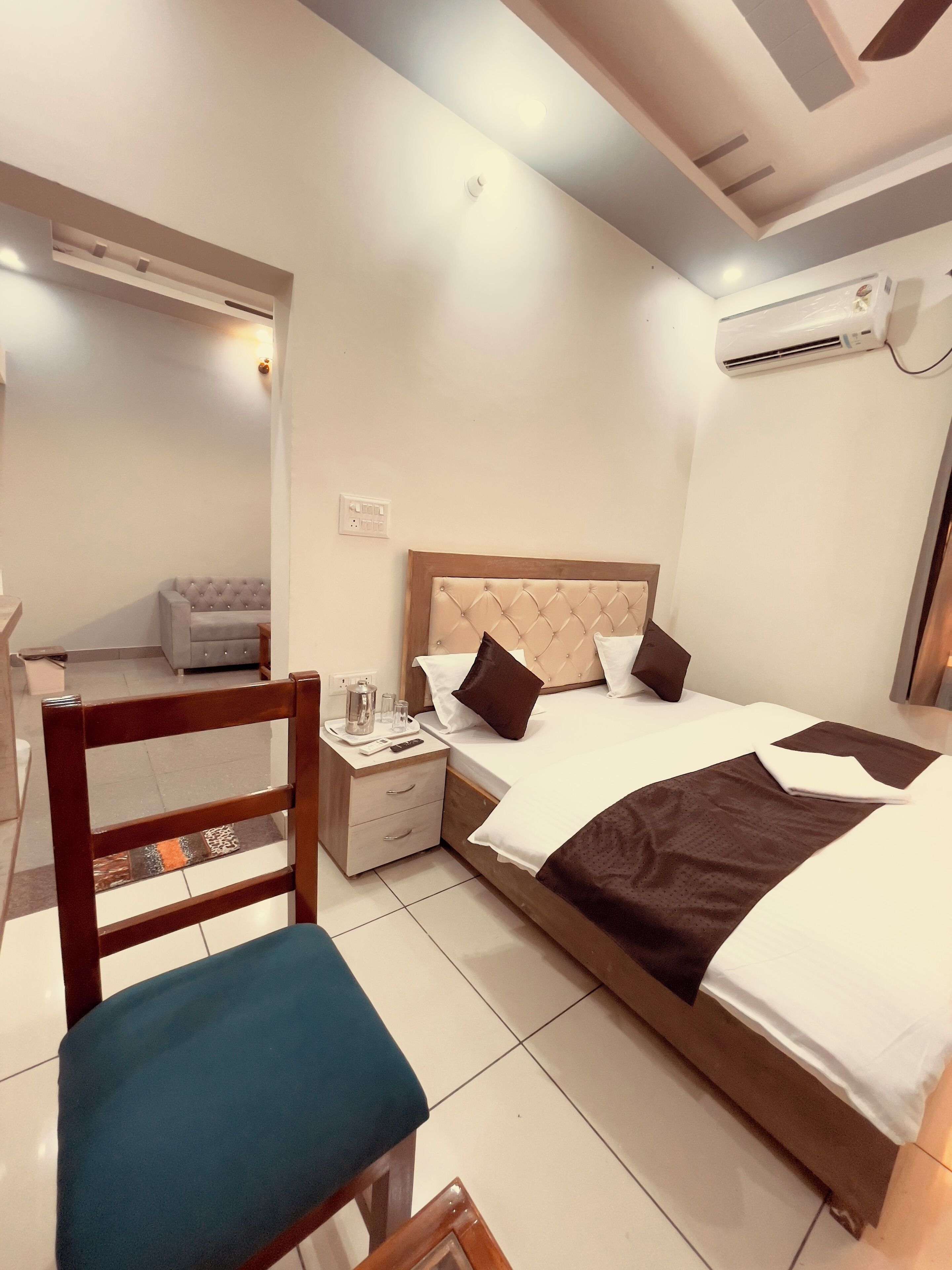 A hotel room with a bed, a wooden nightstand, a chair, a small table, and an air conditioning unit.