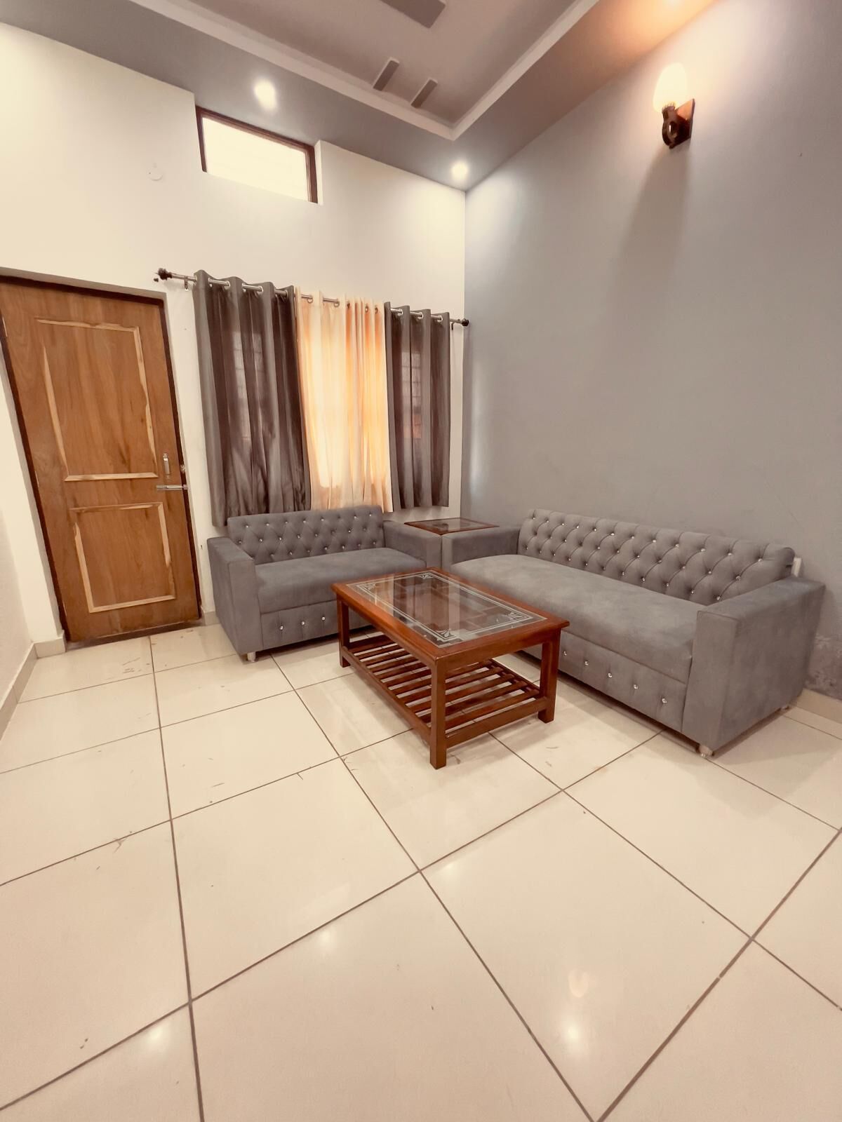 A living room with a sectional sofa, a glass-top coffee table, a wooden door, and a window with curtains.