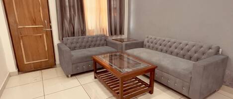 A living room with a sectional sofa, a glass-top coffee table, a wooden door, and a window with curtains.