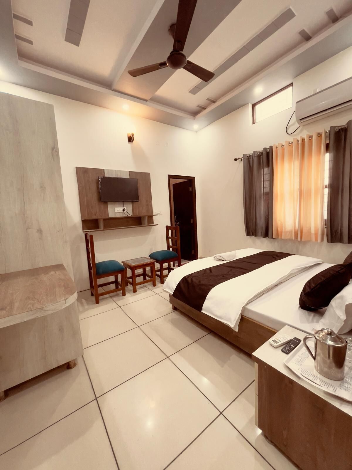 A hotel room with a bed, a desk with chairs, a ceiling fan, and a TV mounted on the wall.