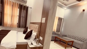 A modern bedroom with a bed, bedside table, and a grey sofa in a living area. - Vishram Guest House (Vikasnagar)