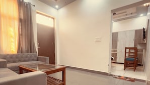 A modern living room with a sofa, a coffee table, and a ceiling fan. There is a door leading to a kitchen area. - Vishram Guest House (Vikasnagar)