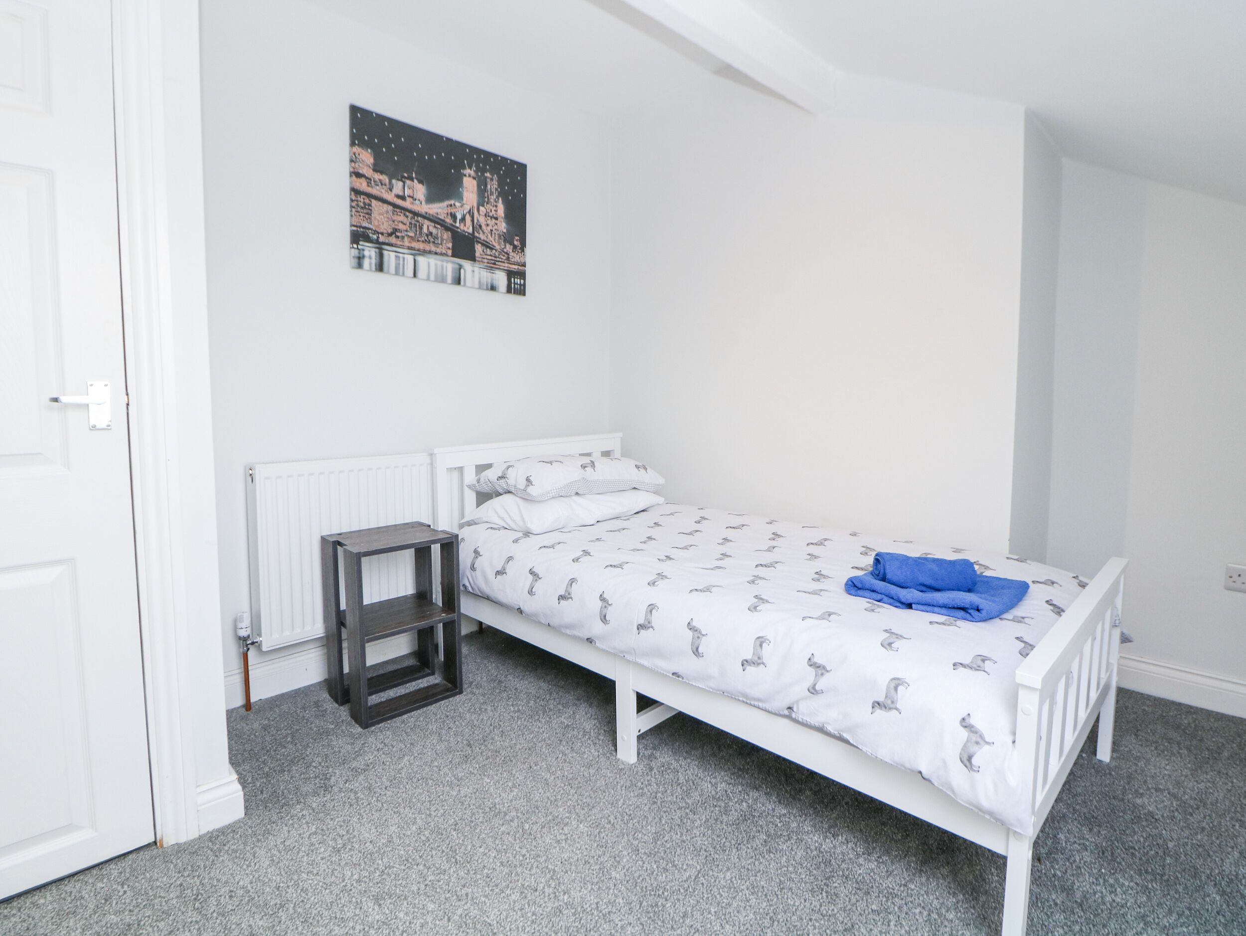 Habitación individual con una cama de matrimonio y cabecera blanca, una mesita de noche pequeña, un radiador y un cuadro en la pared.