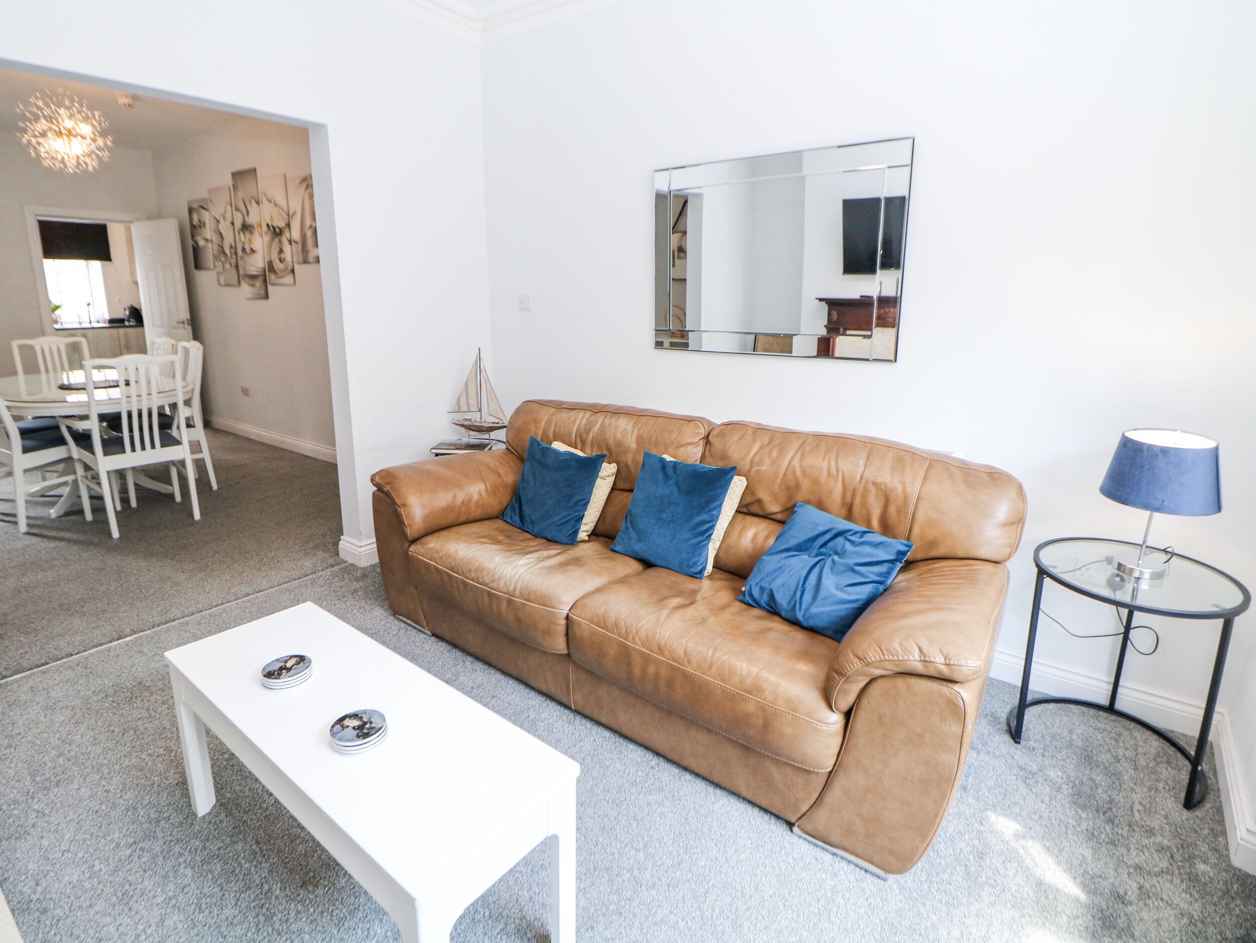 Un salón con un sofá de cuero marrón, dos cojines azules, una mesa de centro blanca, una mesita auxiliar con una lámpara y un espejo grande en la pared.