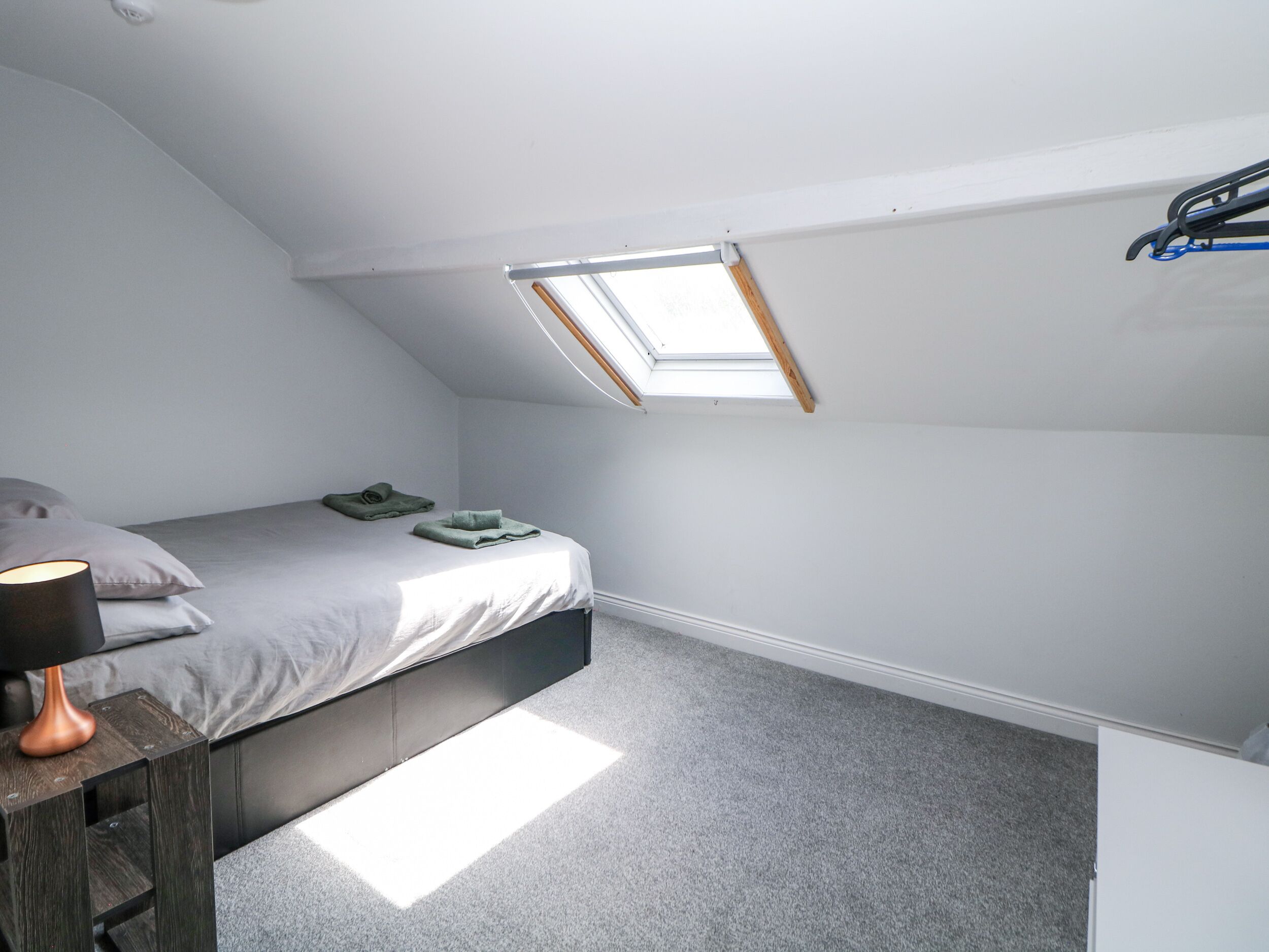 A small, modern bedroom with a skylight, a bed, a bedside table with a lamp, and a coat rack.