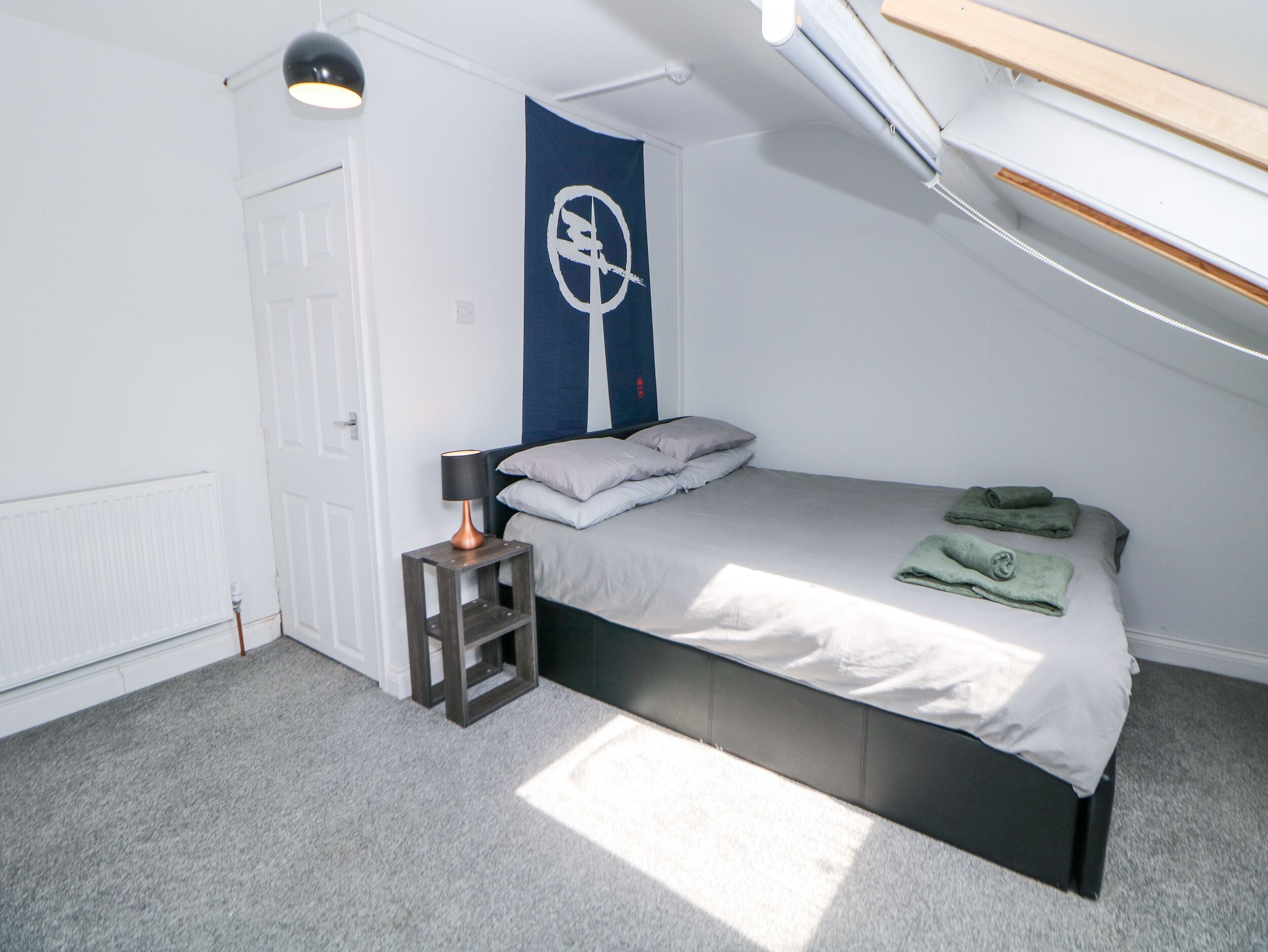 A bedroom with a bed, a nightstand, a lamp, a radiator, and a window.