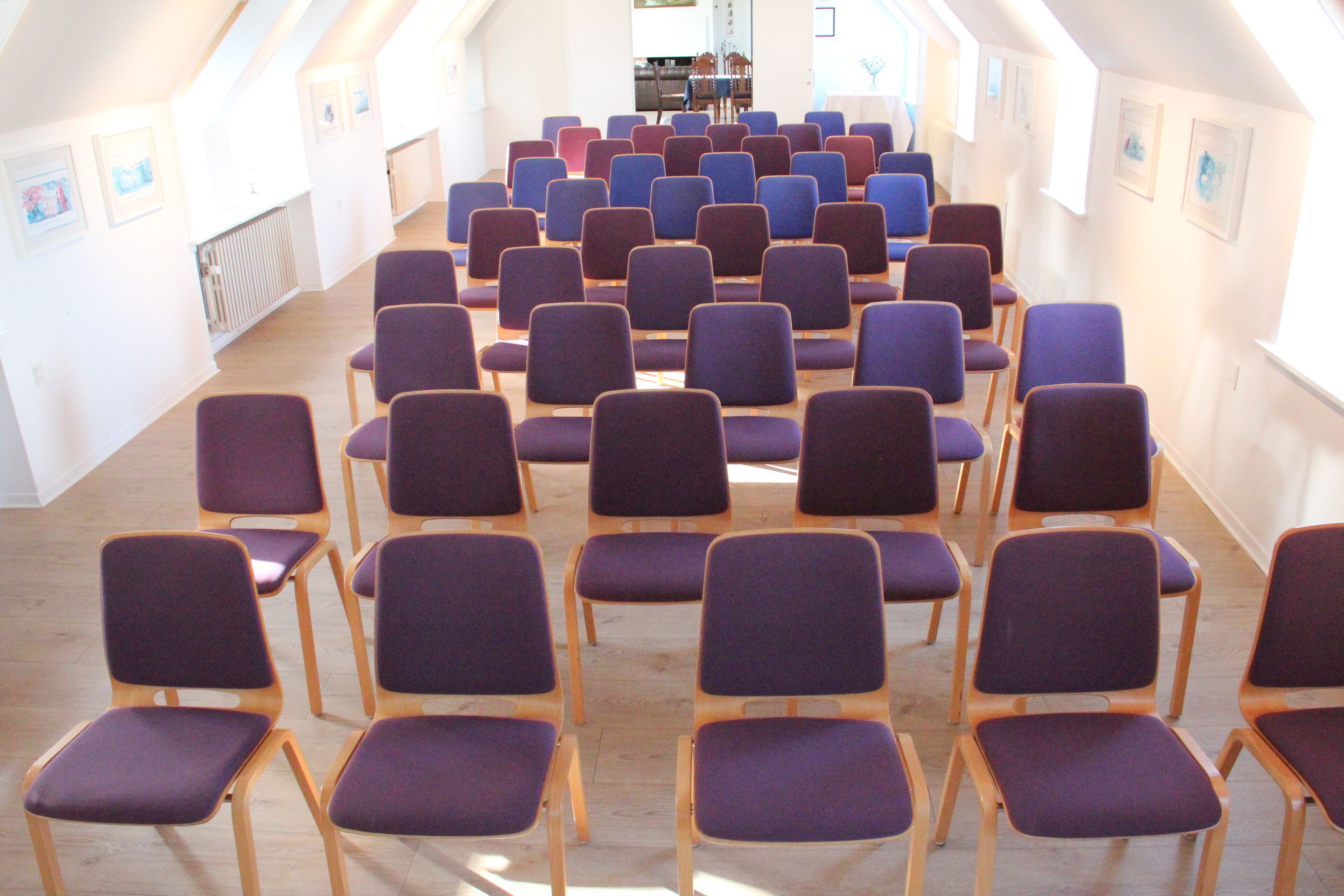 Un salón de conferencias con filas de sillas moradas y azules, piso de madera y paredes blancas con cuadros.