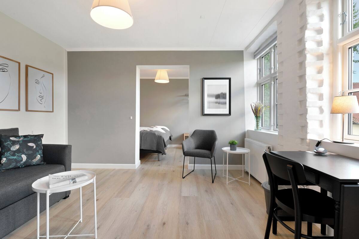 A modern living room with a grey sofa, a round coffee table, a dining table with chairs, and a bed in the background.