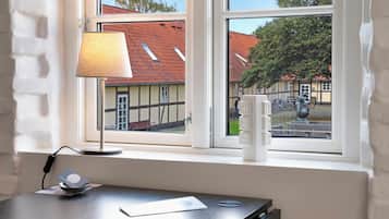 A room with a desk, a lamp, and a window with a view of a building with a red roof.