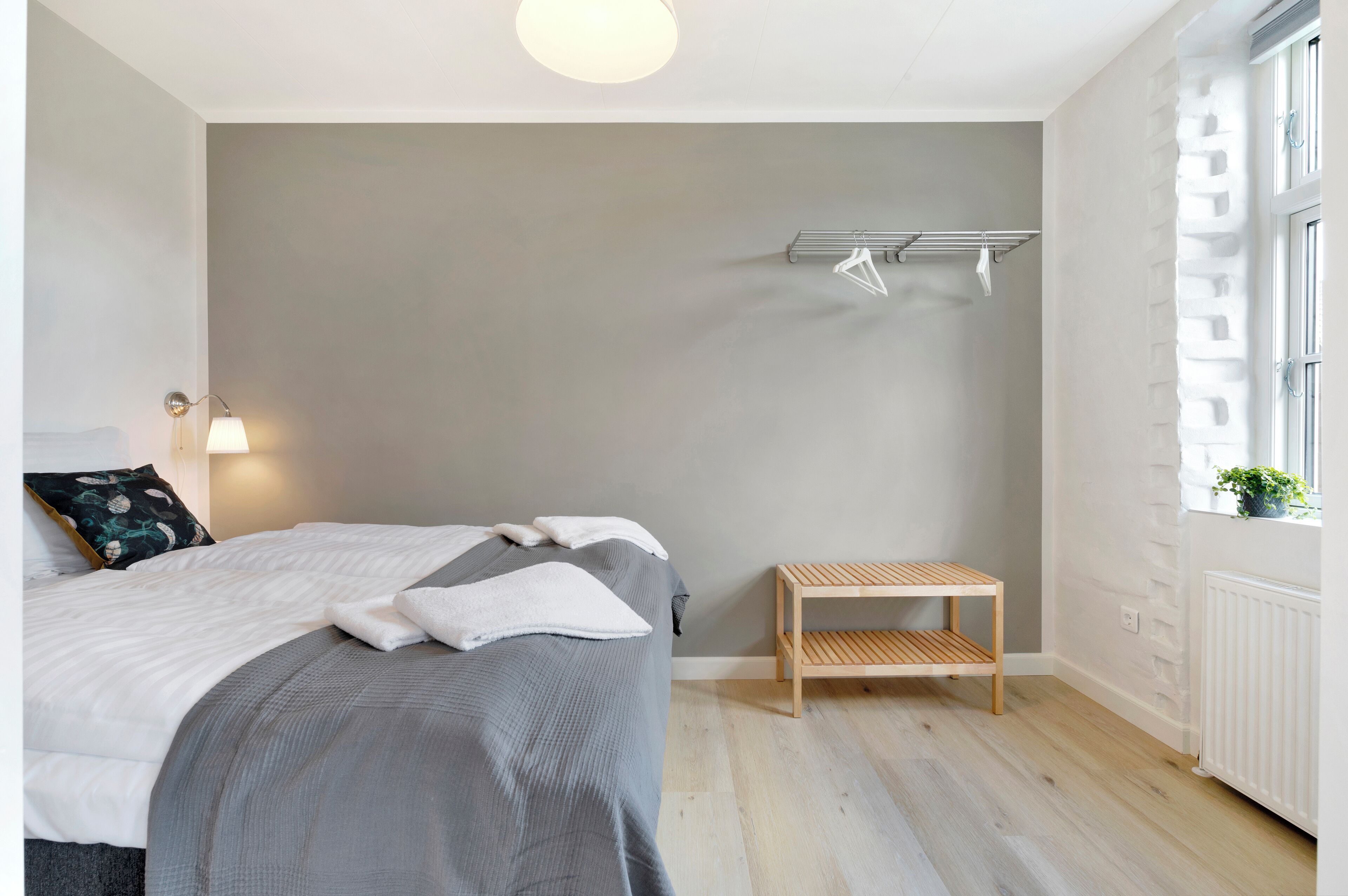 Quarto com cama, mesa de cabeceira, varal fixado na parede e janela com vista.