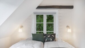A bedroom with a sloped ceiling, a window with a view of trees, two bedside lamps, and a bed with multiple pillows. - Kragsbjerggaard Hotel (Odense)