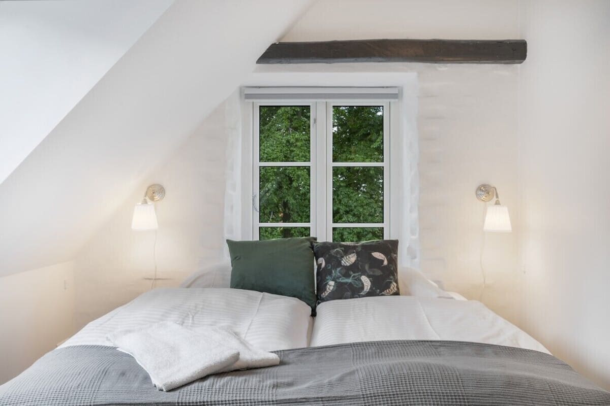 A bedroom with a sloped ceiling, a window with a view of trees, two bedside lamps, and a bed with multiple pillows.