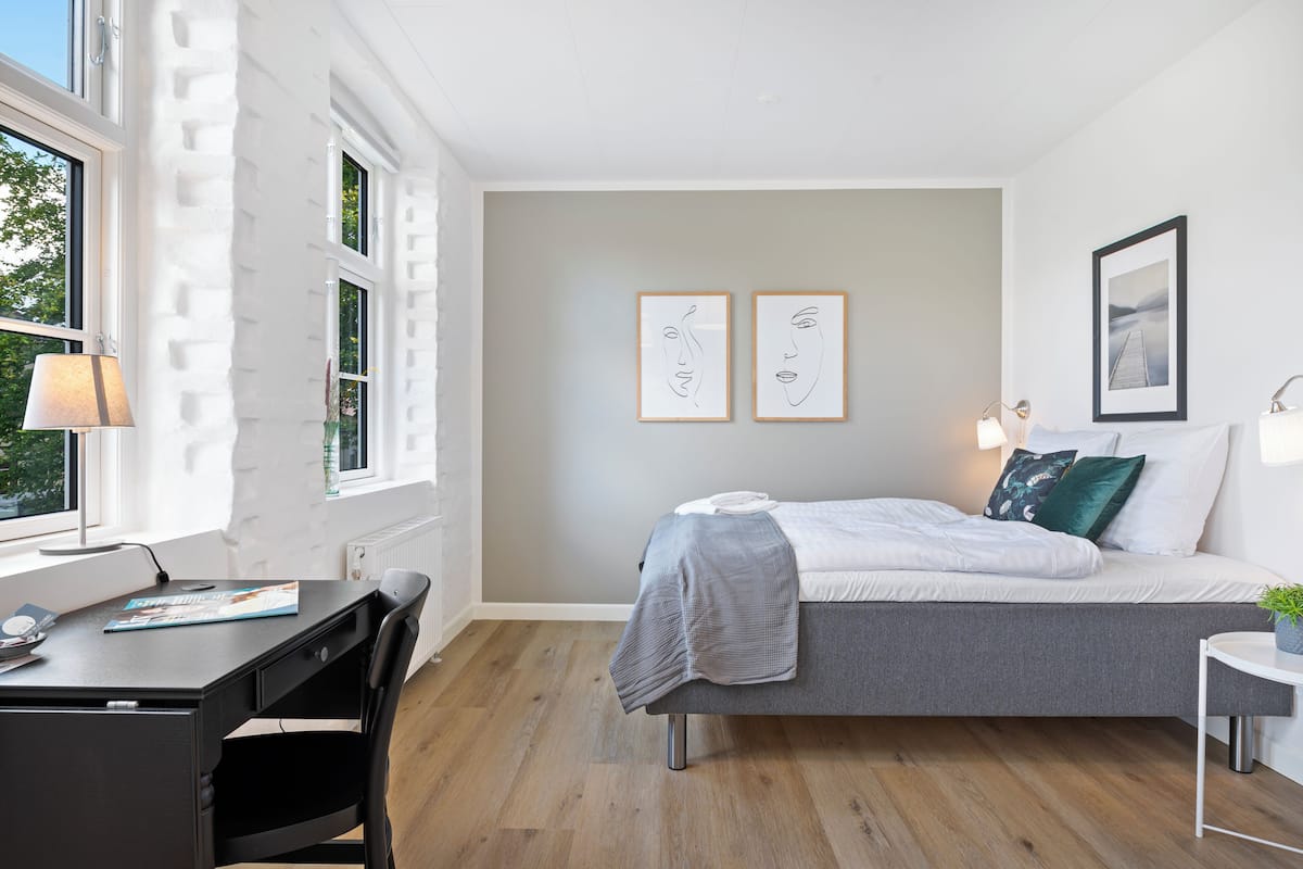 A modern bedroom with a bed, desk, and chair. There are two framed artworks on the wall and a window with a view of trees.