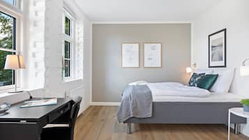 A modern bedroom with a bed, desk, and chair. There are two framed artworks on the wall and a window with a view of trees.