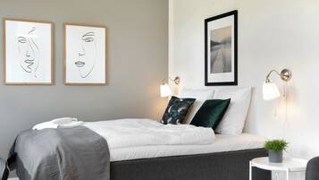 A neatly made bed with white and grey bedding, two wall-mounted picture frames, a small round table with a plant, and two wall-mounted lamps.
