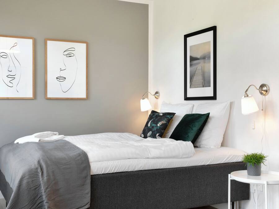 A neatly made bed with white and grey bedding, two wall-mounted picture frames, a small round table with a plant, and two wall-mounted lamps.