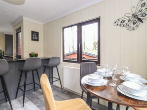 Un comedor con una mesa de madera preparada para cuatro, una cocina al fondo y una ventana con vista a un edificio de techos rojos.