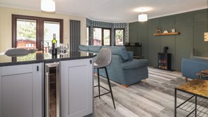 A modern kitchen with a built-in oven, a dining area with a wooden table, a blue sofa, and a TV mounted on the wall. - Alfresco Lodge (Windermere)