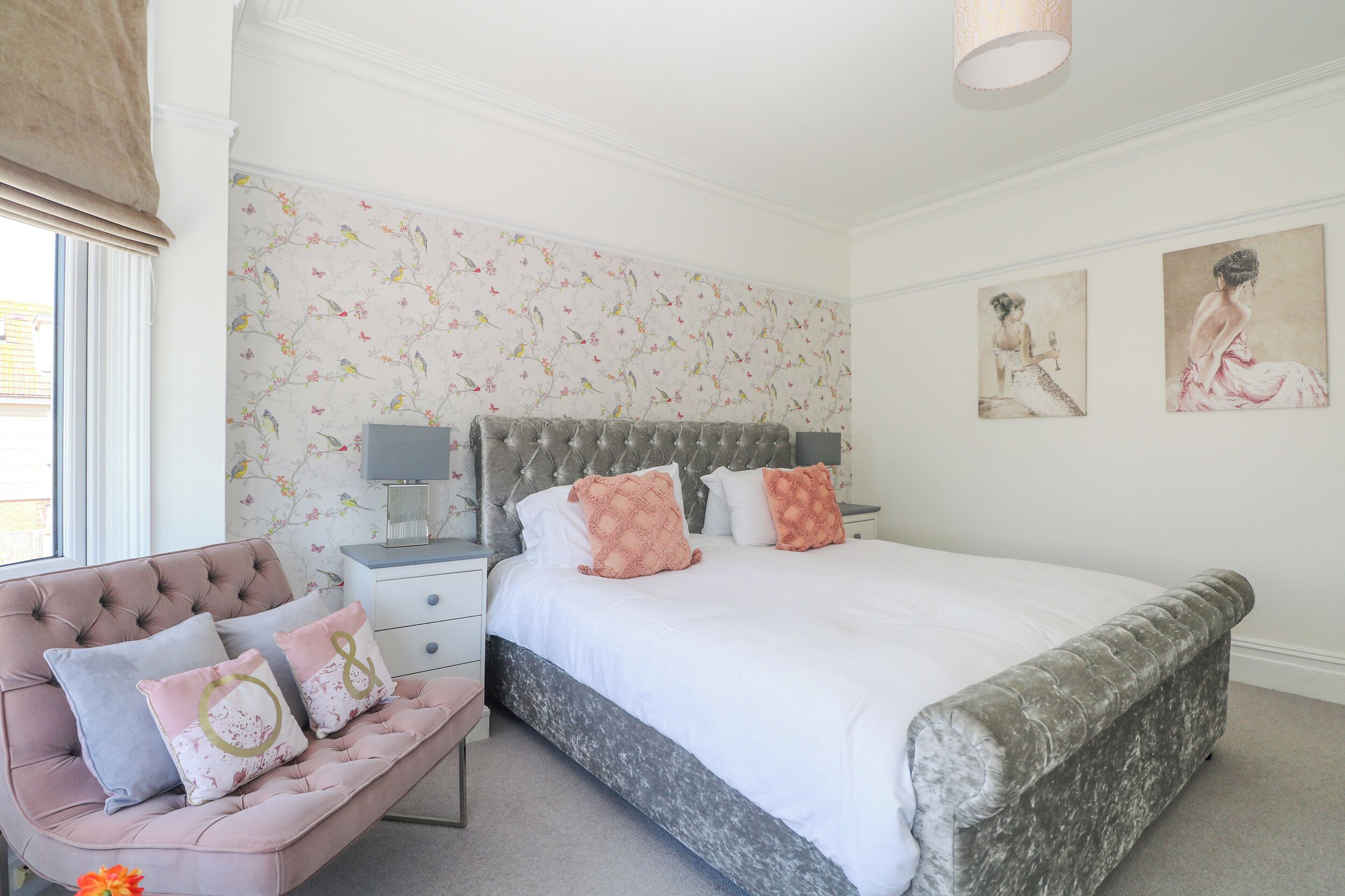 A bedroom with a large bed, a tufted headboard, a bench with cushions, a nightstand, and floral wallpaper.
