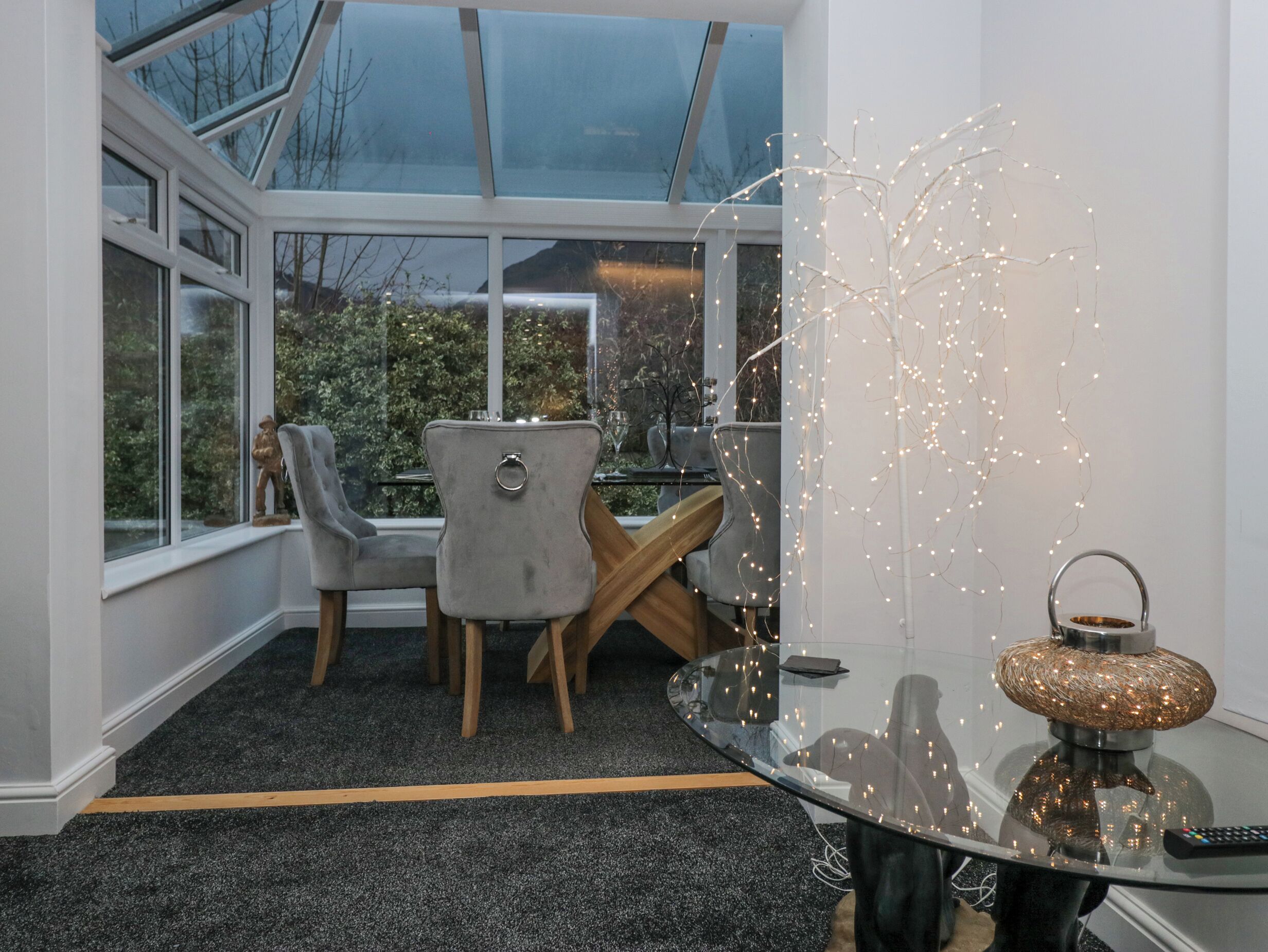 A modern dining area with a glass table, grey upholstered chairs, and a view of greenery outside.