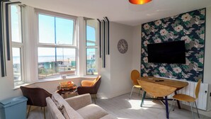 A modern living room with a sofa, chairs, a wooden table, a TV, and large windows with a view. - Apt 4 @ Hunter's Quay (Bridlington)