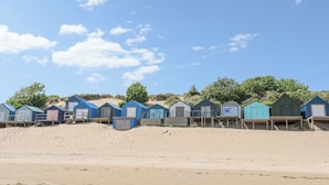 Una hilera de cabañas de playa con pasarelas de madera que las conectan, ubicadas en una playa arenosa bajo un cielo despejado.