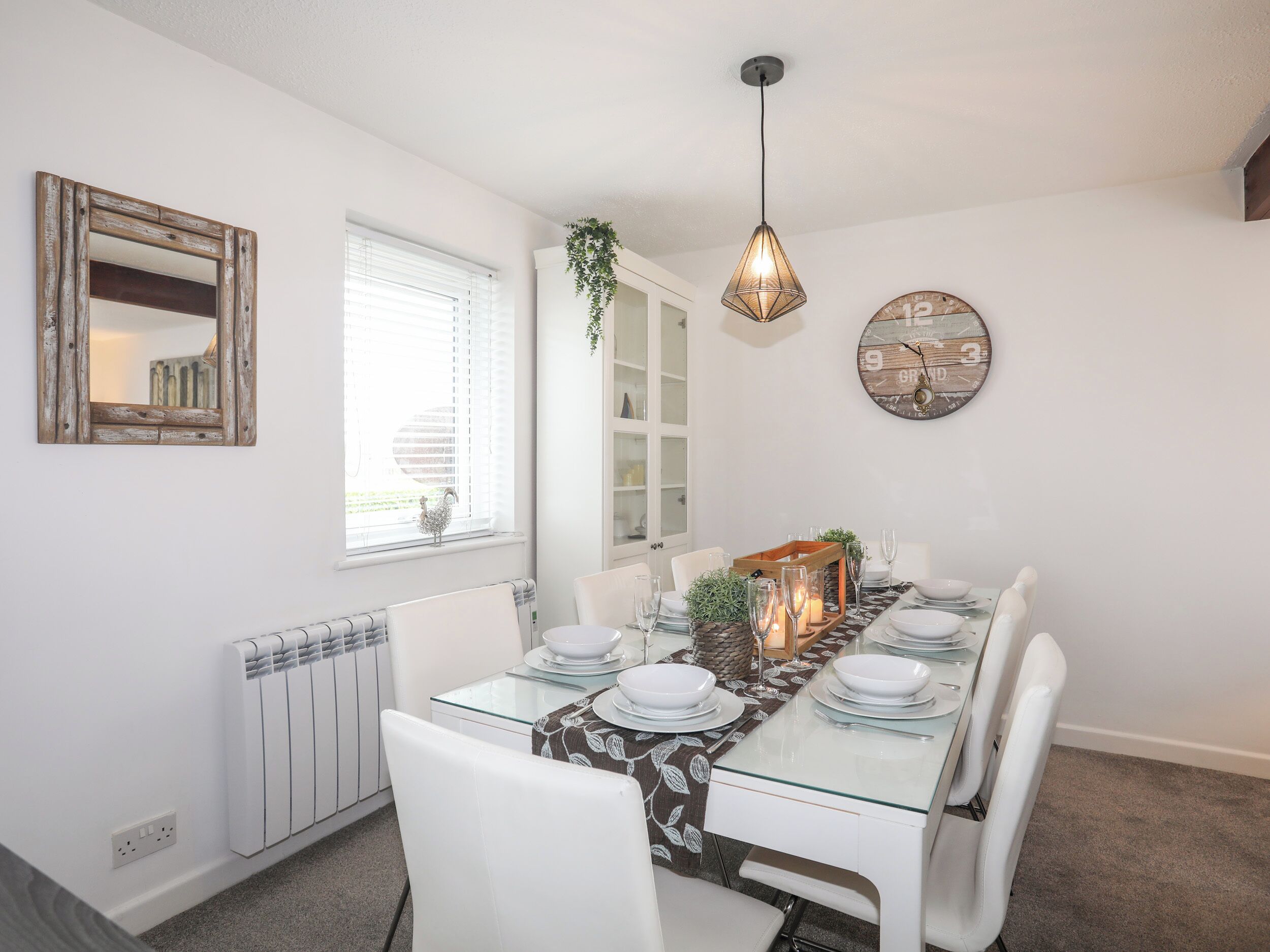 A dining area with a glass table, white chairs, a hanging light fixture, a clock, and a window with blinds.