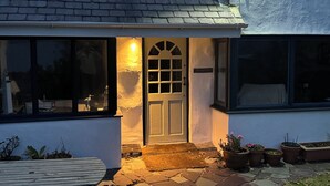 A house with a white exterior, a dark roof, and a front door with a window above it. There are potted plants outside and a bench in the foreground. - Bryn Teg Cottage (Moelfre)