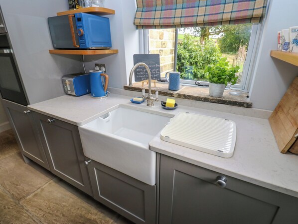 A kitchen with a white sink, a blue kettle, and a window with a view of greenery. - Black House Cottage (Sowerby Bridge)