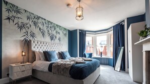 Un dormitorio con cabecera capitonada, una cama grande con ropa de cama azul, una mesita de noche con cajones, una lámpara de pared y una ventana con cortinas.