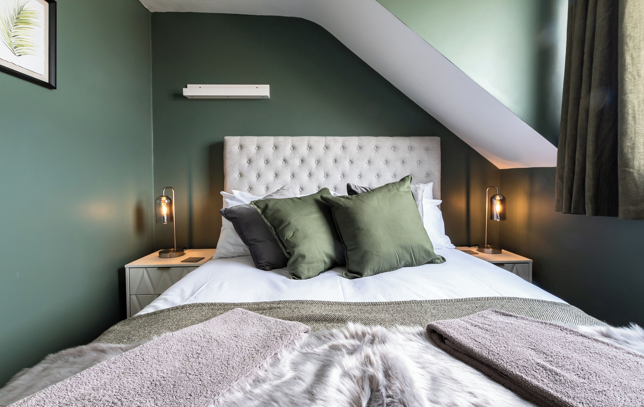 A modern bedroom with a tufted headboard, two bedside tables with lamps, a fluffy throw blanket, and a framed picture on the wall.