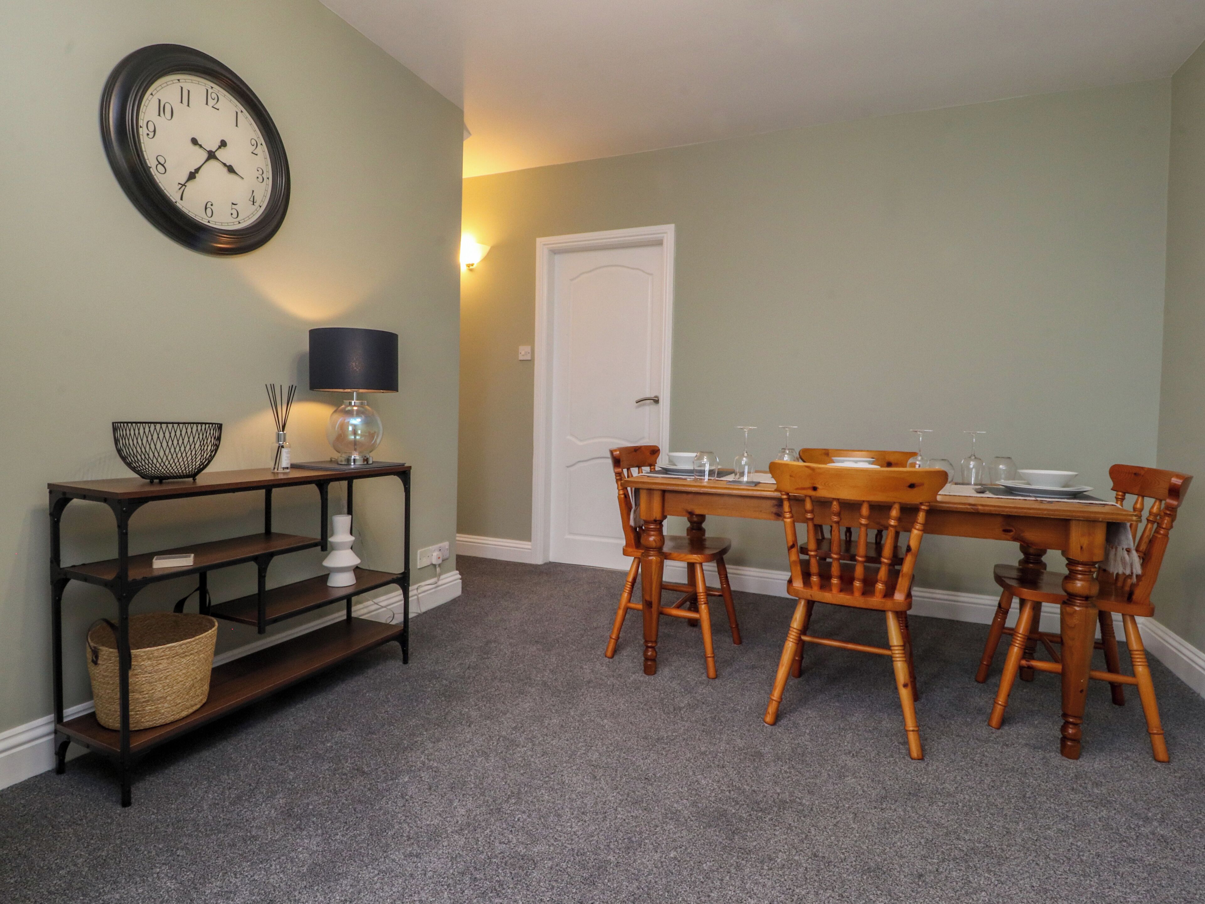 A dining area with a wooden table and chairs, a wall clock, a lamp, and a small shelf with a basket.