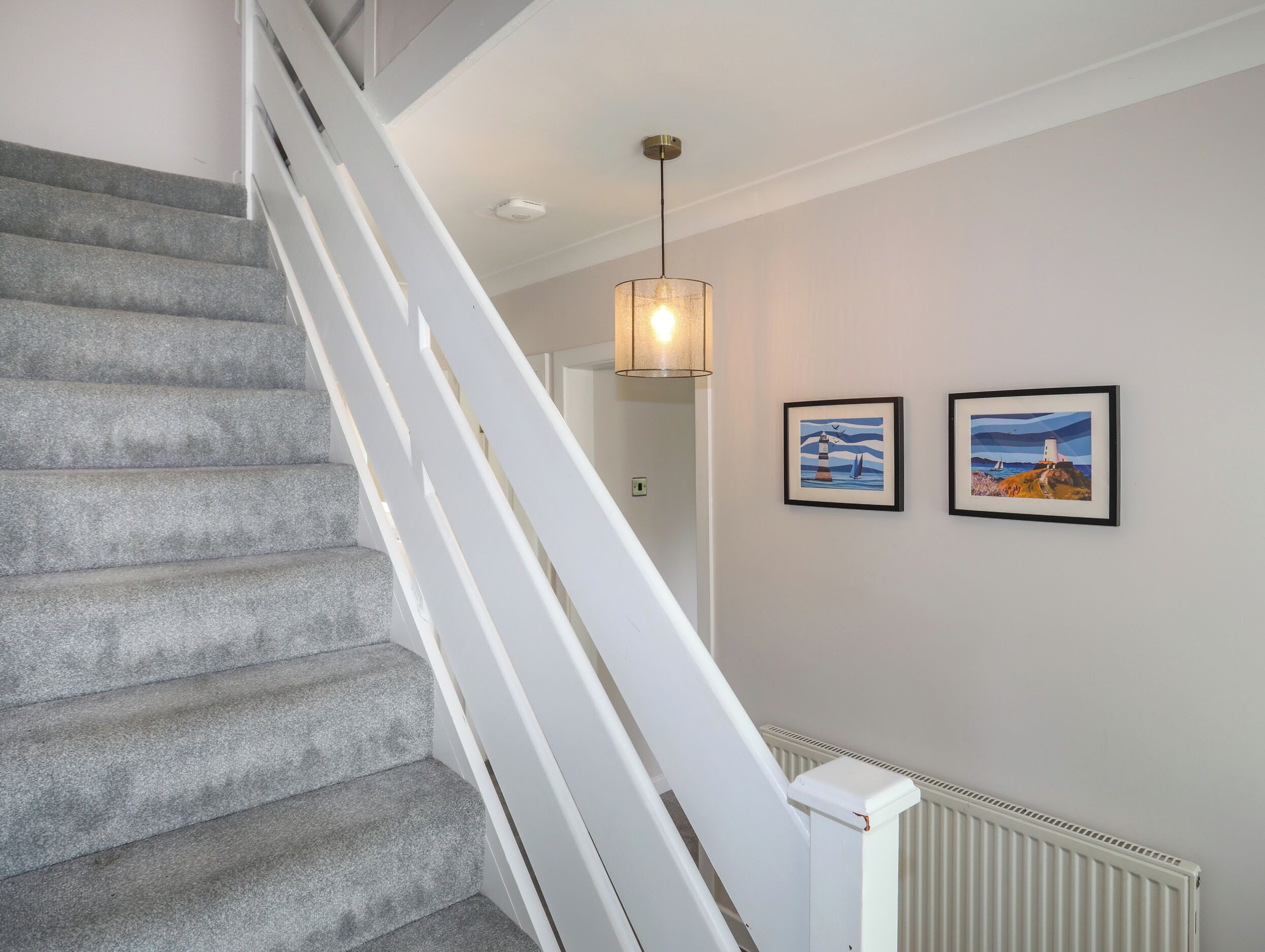 Eine Treppe mit grauen Teppichstufen und weißen Geländern, die zu einem Raum führt, in dem ein weißer Heizkörper und zwei gerahmte Bilder an der Wand hängen.