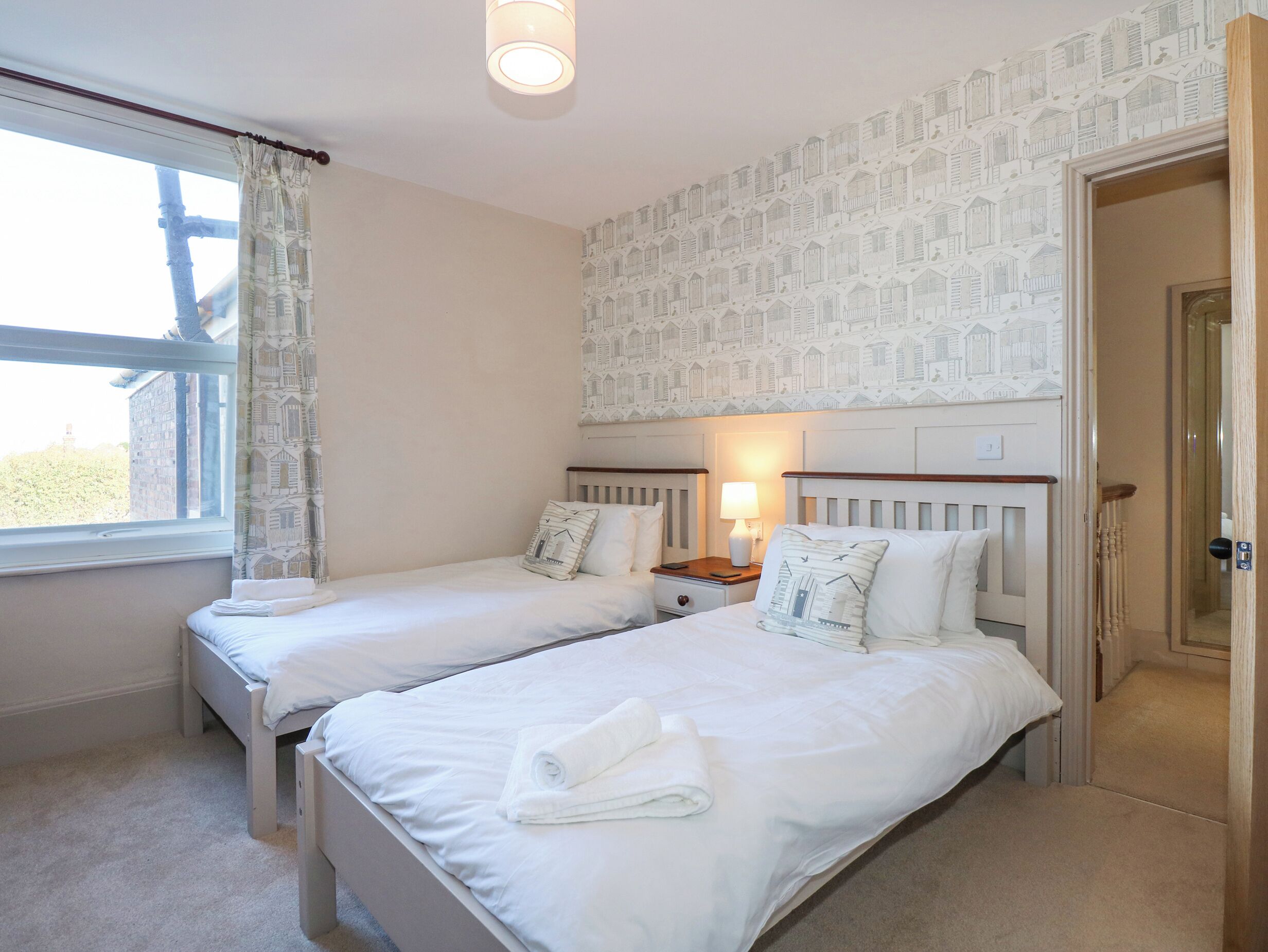 Habitación de hotel con dos camas individuales, una ventana con cortinas, una pared con un diseño de casas de pájaros y una puerta que da a otra habitación.