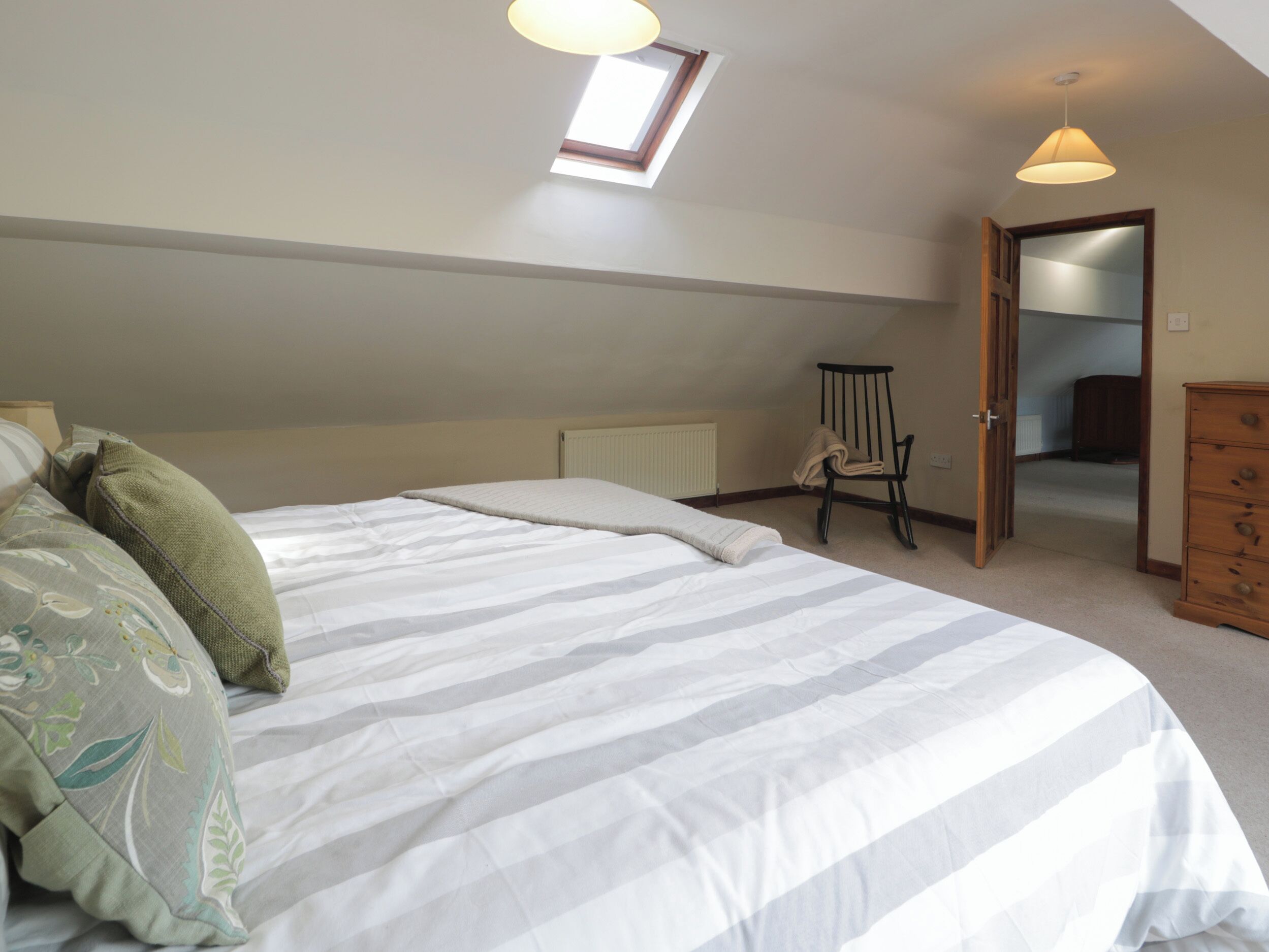 A bedroom with a large bed, a skylight, a chair, and a wooden dresser.