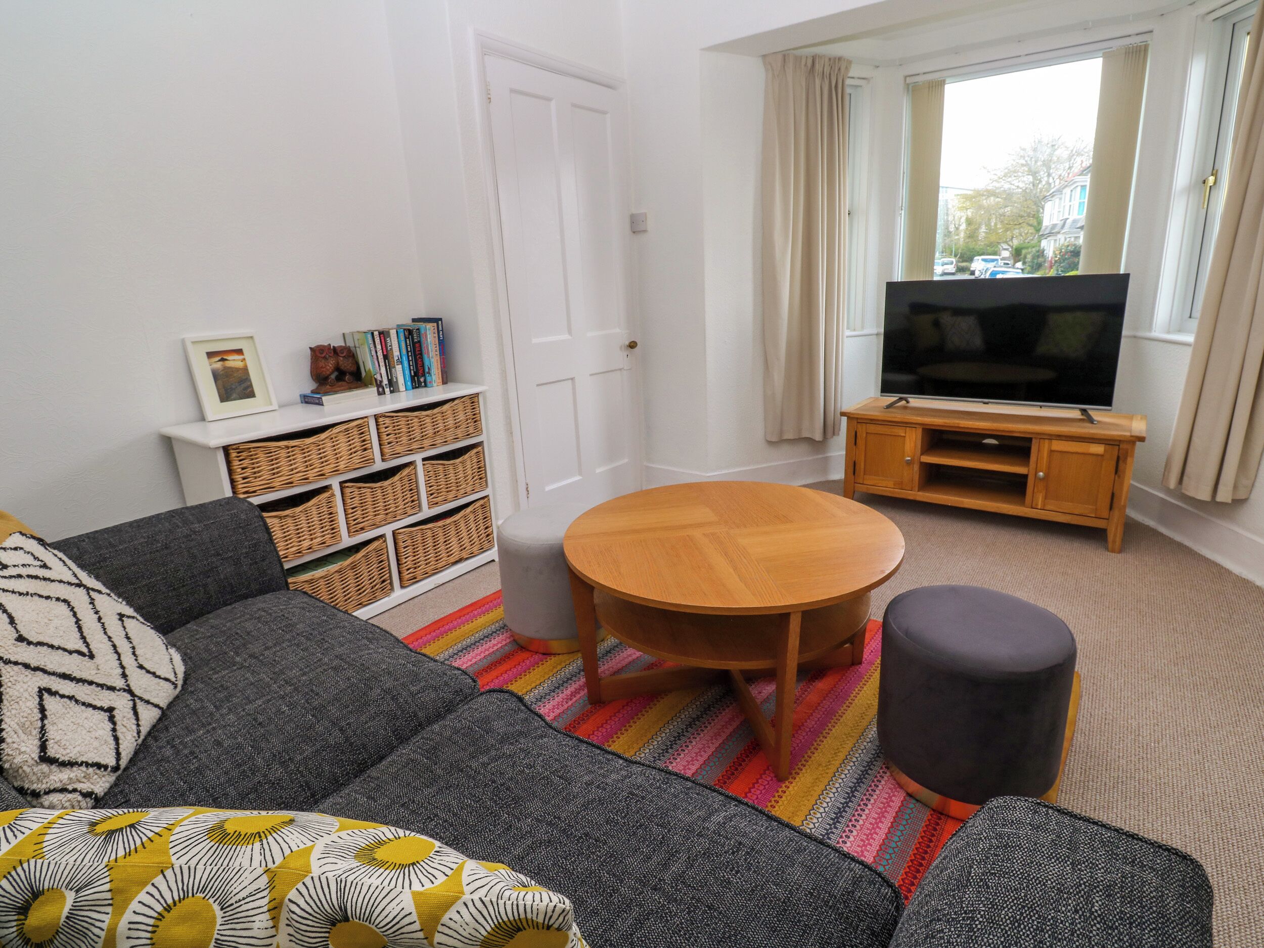 A living room with a sofa, a round wooden table, a television on a wooden stand, and a bookshelf with wicker baskets.