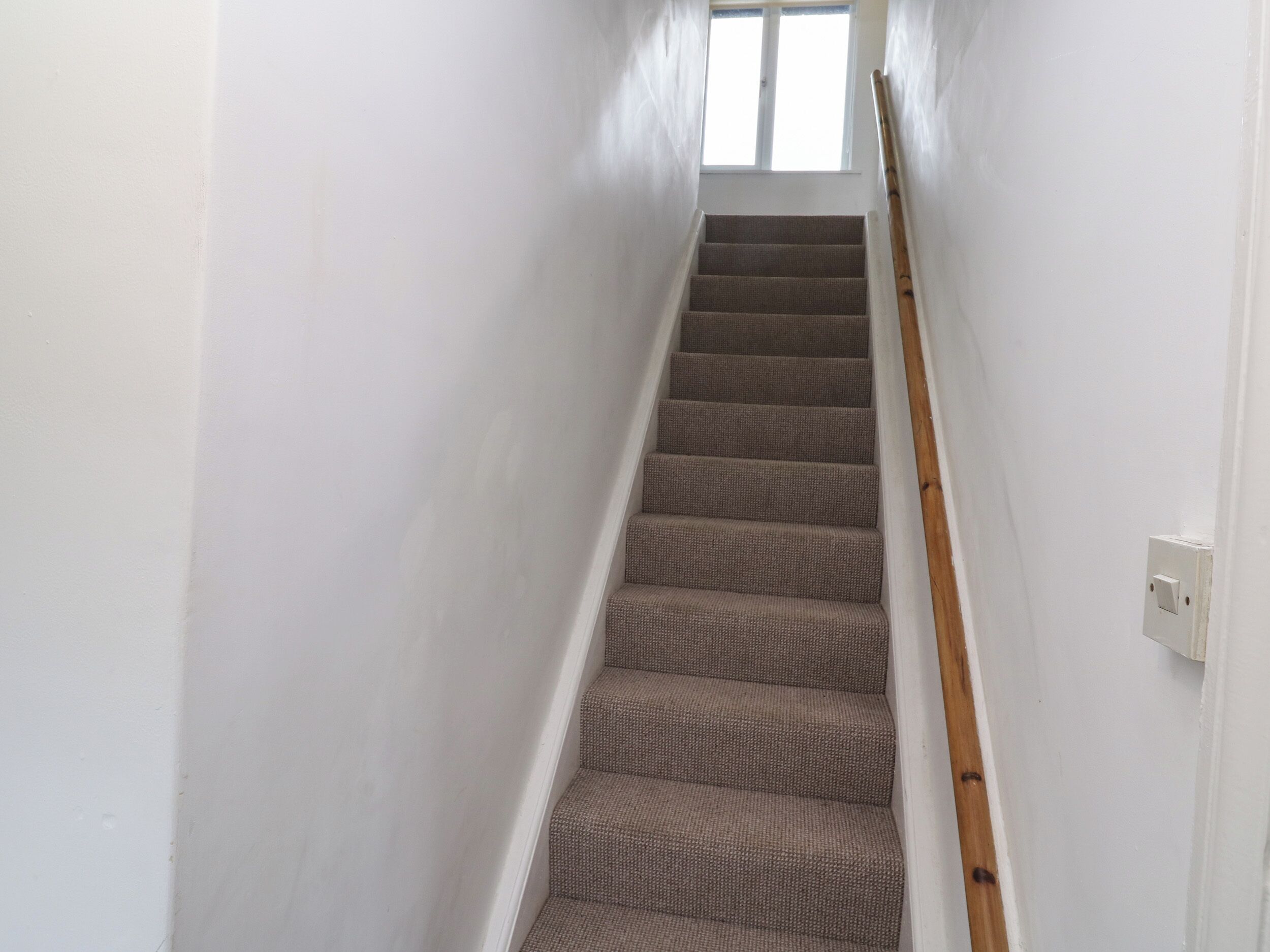 Un escalier étroit, avec des marches recouvertes de moquette et une rampe en bois, menant à une fenêtre située en haut.