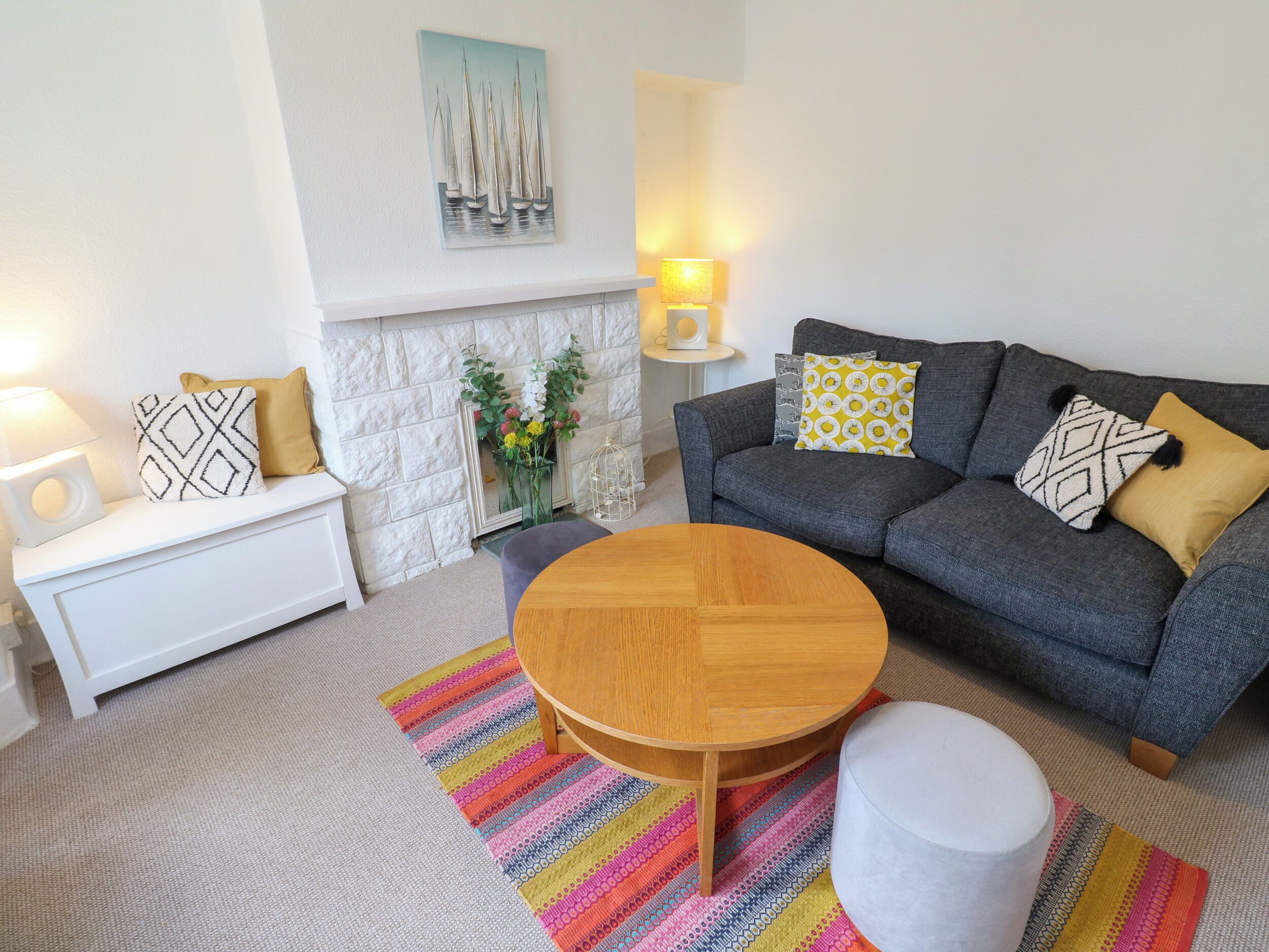 A living room with a grey sofa, a white side table, a round wooden coffee table, a colorful striped rug, and a white ottoman.