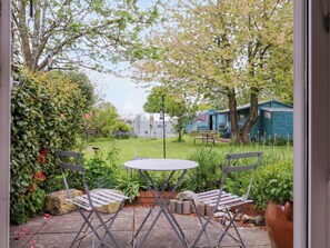 A patio with a round table and two chairs, surrounded by greenery and a view of a garden and buildings in the background.