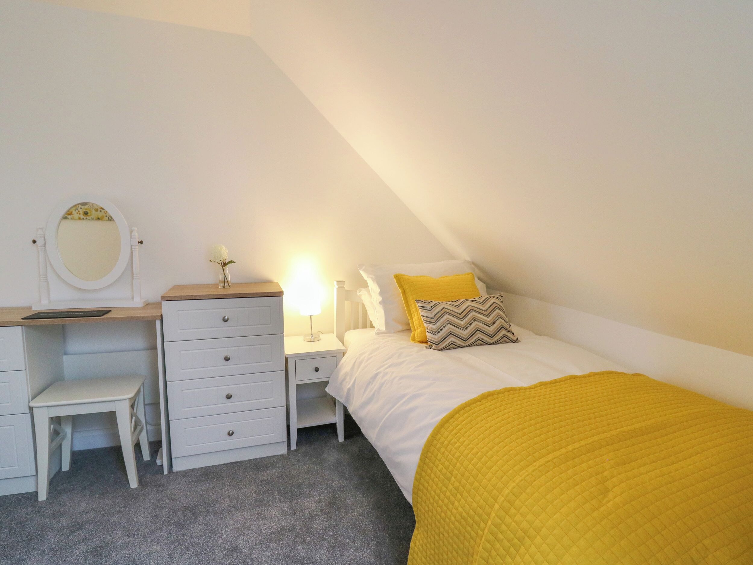 Ein Schlafzimmer mit Bett, Schreibtisch mit Schubladen, Spiegel, Nachttisch und Lampe.