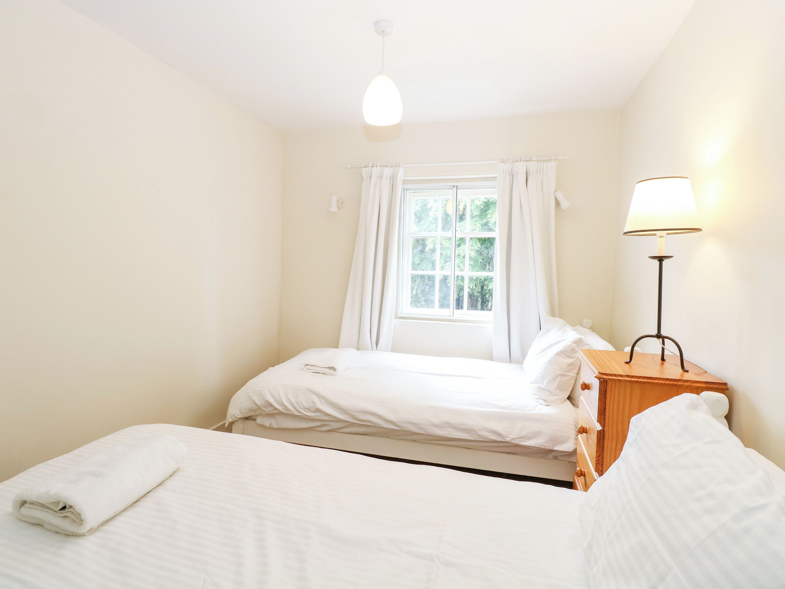 Un dormitorio con dos camas, una ventana con cortinas, una mesita de noche con lámpara e iluminación en la pared.