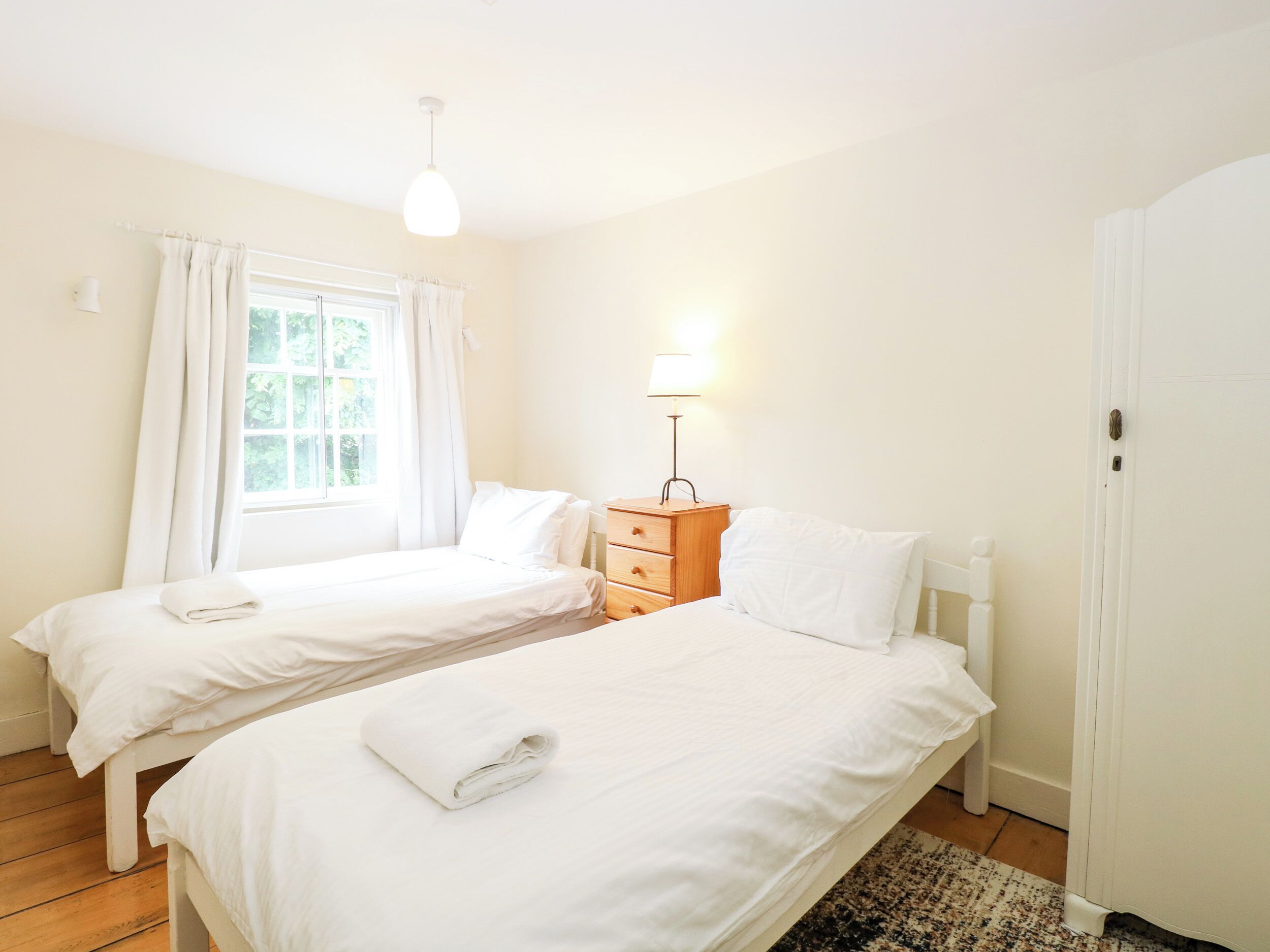A room with two beds, a wooden nightstand with a lamp, a window with curtains, and a door.