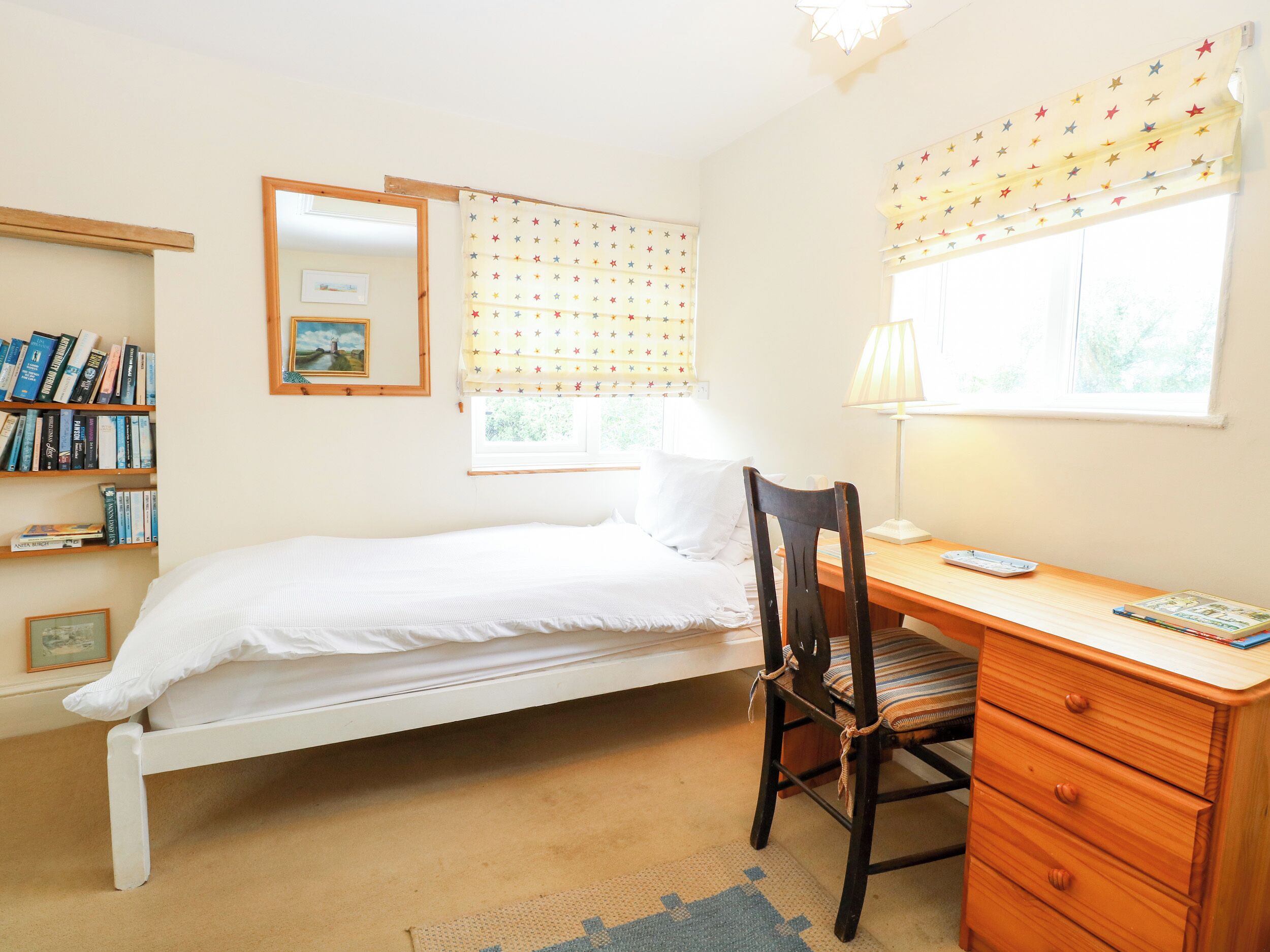 Slaapkamer met een enkel bed, een houten bureau met stoel, een boekenkast en een raam met gordijnen met stero patroon.