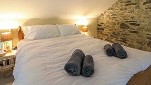 Un lit bien fait, avec du linge de lit blanc et deux serviettes enroulées au centre. Il y a deux lampes de chevet avec des abat-jour, et une pile de livres sur la table de chevet. Un mur en pierre orne un des côtés de la pièce.