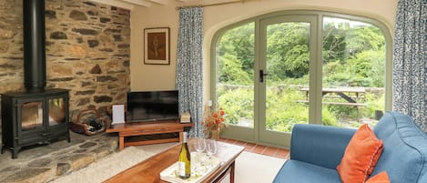 A cozy living room with a stone fireplace, a television, a sofa, and a large window offering a view of greenery.