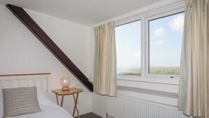 A bedroom with a sloped ceiling, a bed with a striped pillow, a bedside table with a lamp, and a window with curtains. - Golygfa Rhiw (Pwllheli)