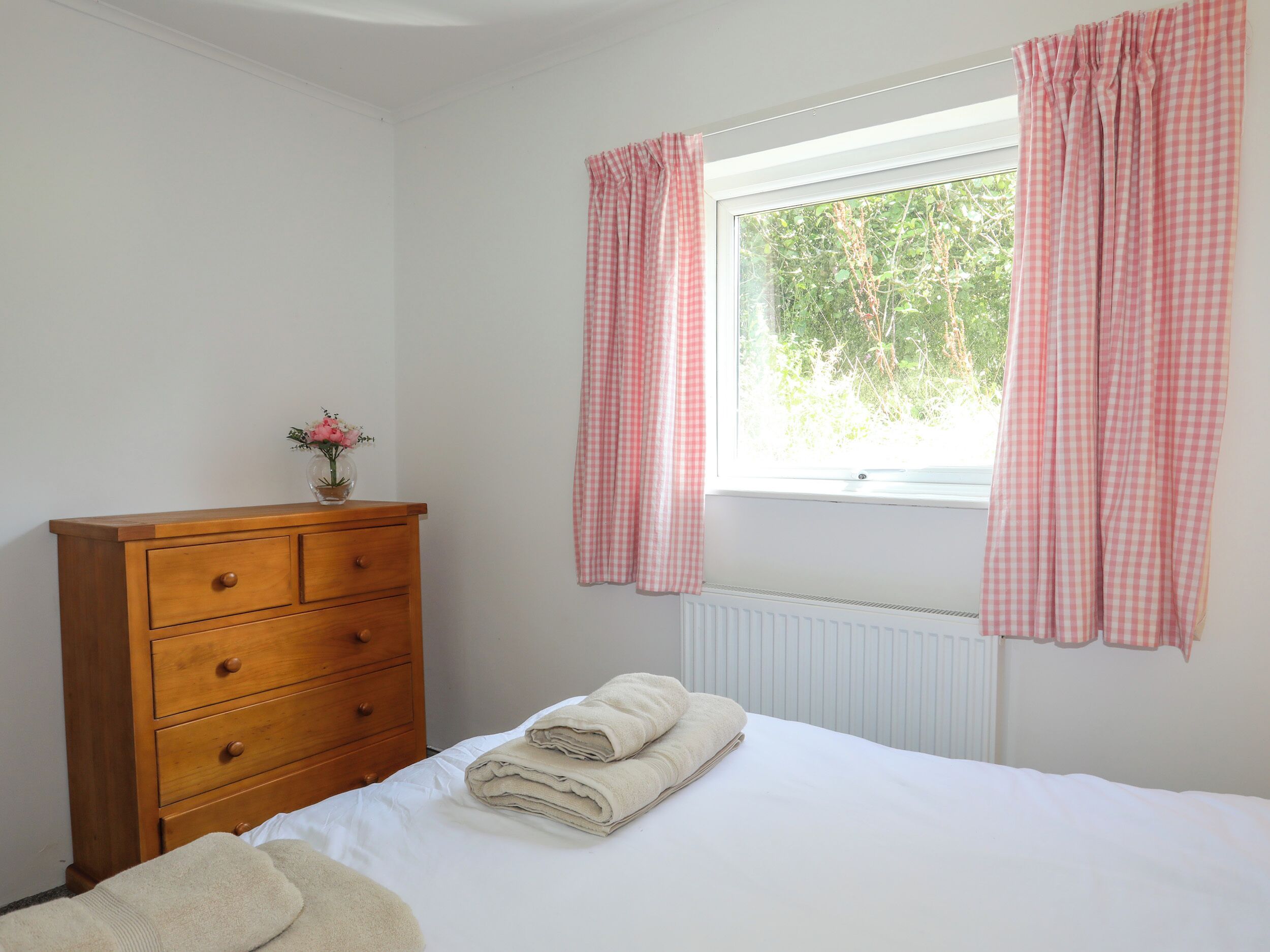 A bedroom with a wooden dresser, a window with pink checkered curtains, and a radiator.
