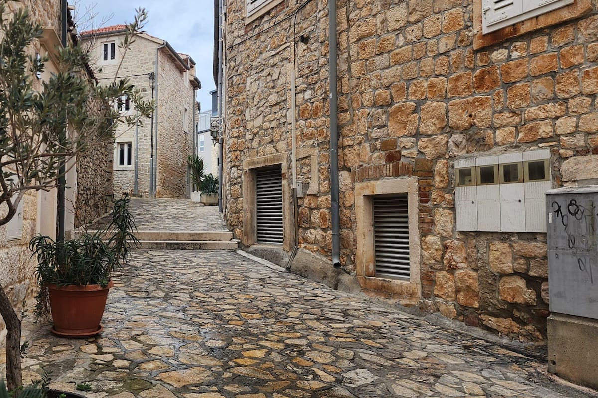 A narrow cobblestone street flanked by stone buildings with shutters and a potted plant.